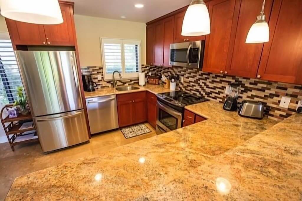 The kitchen features modern stainless steel appliances, including a refrigerator and a dishwasher, set against rich wood cabinetry. A textured granite countertop spans the work area, complemented by a decorative backsplash. Natural light enters through a nearby window, illuminating the inviting space.