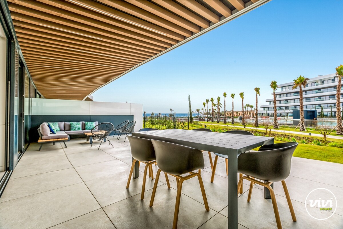 A spacious terrace features a dining table with chairs and a lounge area with cushions. Surrounding greenery is complemented by palm trees, providing a view of the horizon where the sky meets the sea.