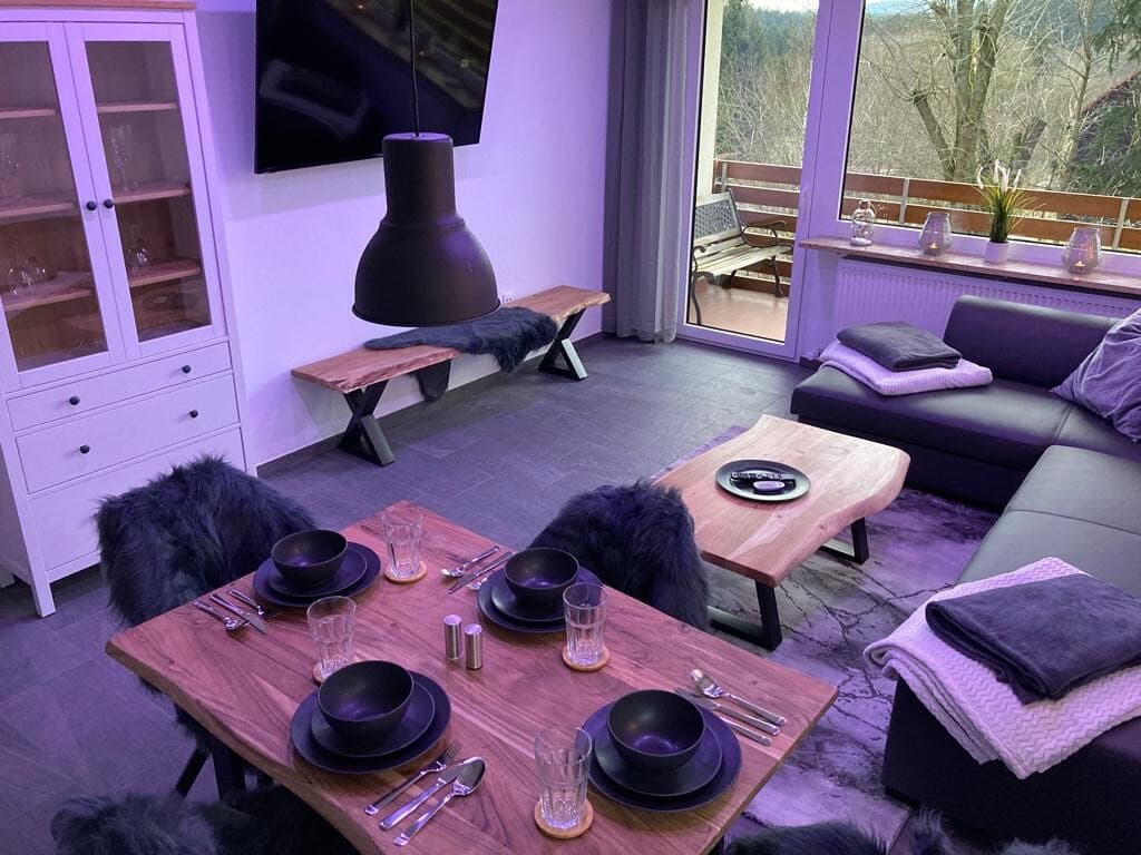 A modern living area features a stylish dining table set for four, accompanied by dark tableware and glassware. A cozy seating arrangement includes a sectional sofa and a low, rustic coffee table. Large windows showcase a view of the balcony and surrounding greenery.