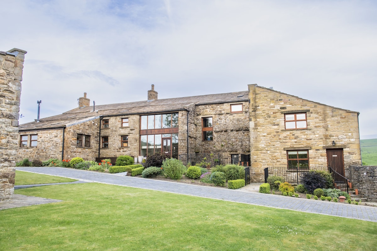 The stone exterior of Hill Top Farm exhibits traditional architecture, complemented by large windows that invite natural light. A well-manicured lawn is visible in the foreground, featuring landscaped gardens that create a welcoming entrance to the property.