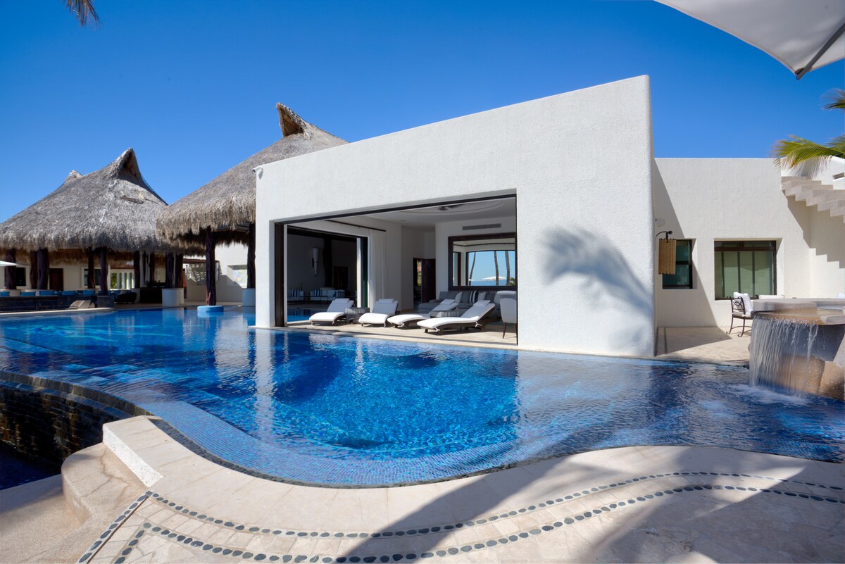 An elegant villa exterior is seen with a spacious terrace overlooking a large infinity pool. Sun loungers are arranged nearby under a shaded area. The architectural design features open spaces and large windows, allowing for ample natural light and stunning views of the surrounding landscape.