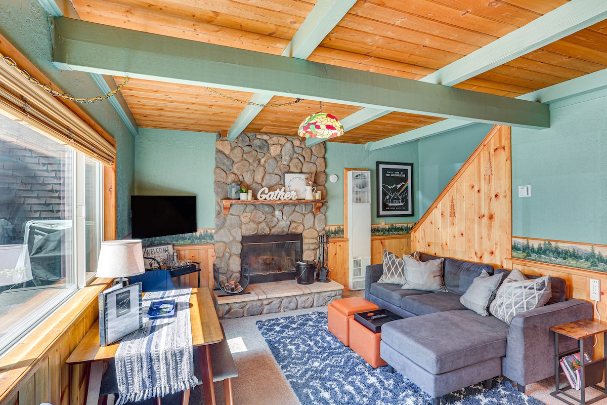A living room features wooden beams on the ceiling, a stone fireplace surrounded by wood paneling, and a large sectional sofa. Natural light floods in through a window, and a coffee table stands in front of the sofa, creating a welcoming space for relaxation.