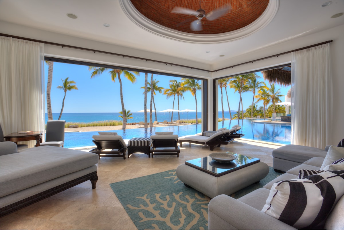 The open living area features large glass doors that extend to a terrace, offering unobstructed views of the ocean and palm trees. A patterned rug lies on a tiled floor, complemented by modern furnishings, including a round coffee table and plush sofas, inviting relaxation.
