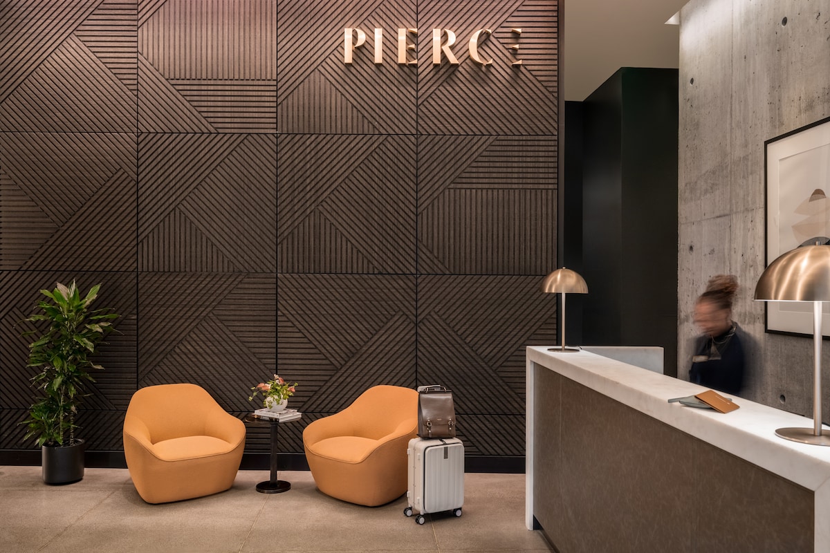The welcoming lobby features a textured wall with the name 'PIERC' elegantly displayed. Two modern orange chairs are positioned near a small table adorned with fresh flowers. A light-colored reception desk stands nearby, complemented by contemporary floor lamps. A suitcase indicates a guest's presence.
