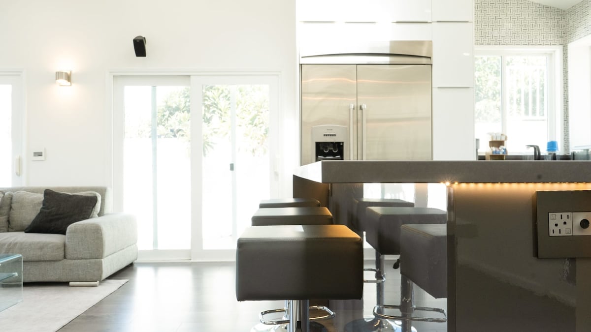 A modern living area featuring a spacious grey sectional sofa next to a glass coffee table. Countertop seating with four black stools creates a casual dining space, while large sliding glass doors illuminate the room and provide access to the outside.