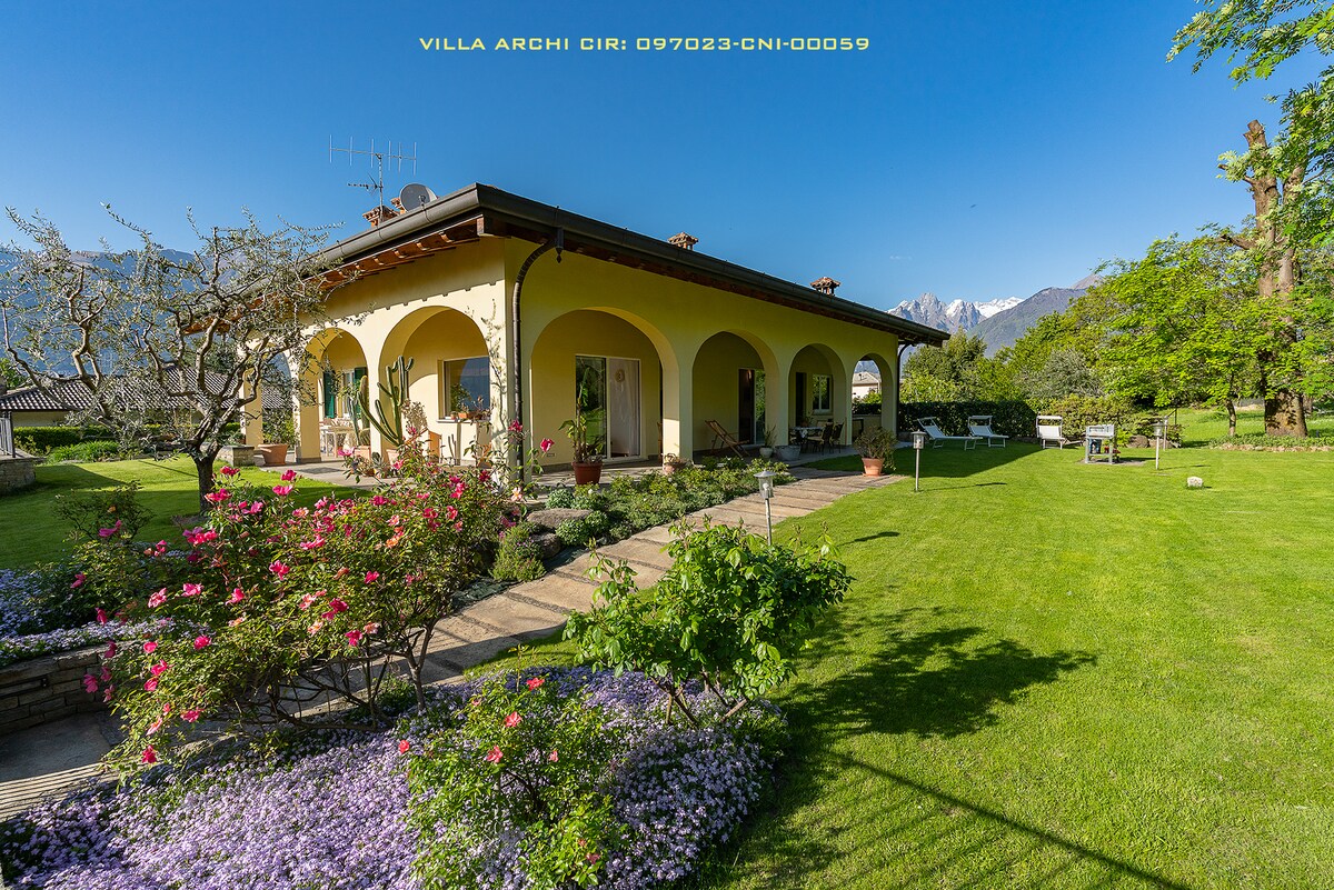 The villa is surrounded by a spacious garden filled with colorful flowers, featuring a stone pathway leading to the covered terrace. Arched windows and a yellow facade enhance the modern design of the home, with mountains visible in the background.
