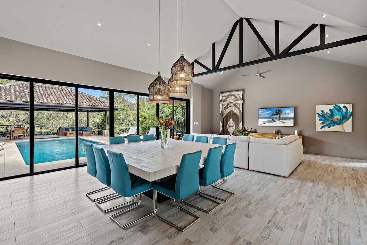 An open-plan living area showcases a large dining table surrounded by light blue chairs. Large glass doors provide a view of the pool area outside, and natural light fills the space. Decorative ceiling fixtures hang above the dining table, adding an elegant touch.