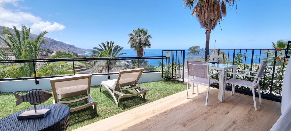 A private terrace is displayed featuring two lounge chairs and a small dining set. The grassy area and palm trees frame the view of the ocean and surrounding hills under a clear blue sky.