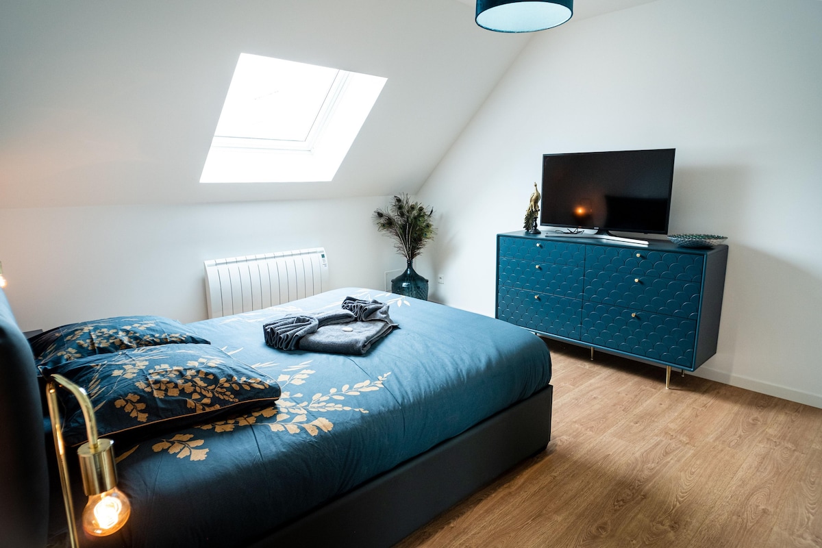 A well-appointed bedroom features a spacious bed with a dark blue coverlet adorned with gold leaf patterns. A sleek television is positioned on a stylish blue dresser, while a skylight allows natural light to fill the room. Decorative elements include a plant and a bedside lamp.