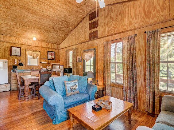 The living room area features a blue sofa and a light wood coffee table. A second couch is positioned nearby. Large windows allow natural light to fill the room, accented by patterned curtains. A dining area with a wooden table and chairs is visible in the background.