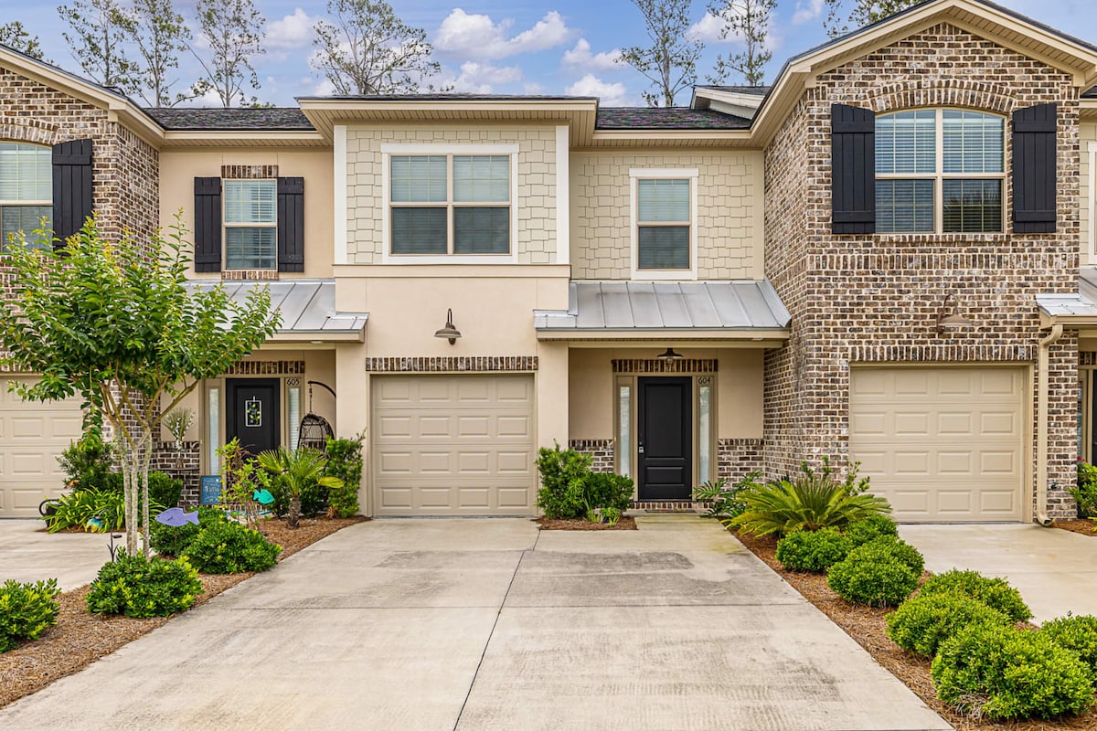 A row of townhomes features a blend of brick and textured siding. Each unit has a welcoming front porch area, flanked by neatly trimmed landscaping. A driveway leads to a single-car garage for each home, providing a structured and cohesive community appearance.
