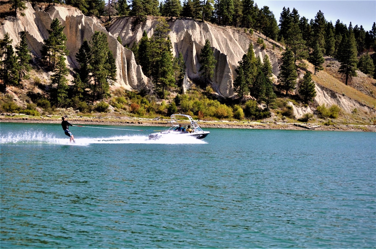A person is seen water skiing beside a boat on a clear blue lake. Lush green trees line the shoreline, providing a backdrop to the sandy cliffs rising in the distance. The surface of the water reflects the surrounding landscape, creating a serene atmosphere.
