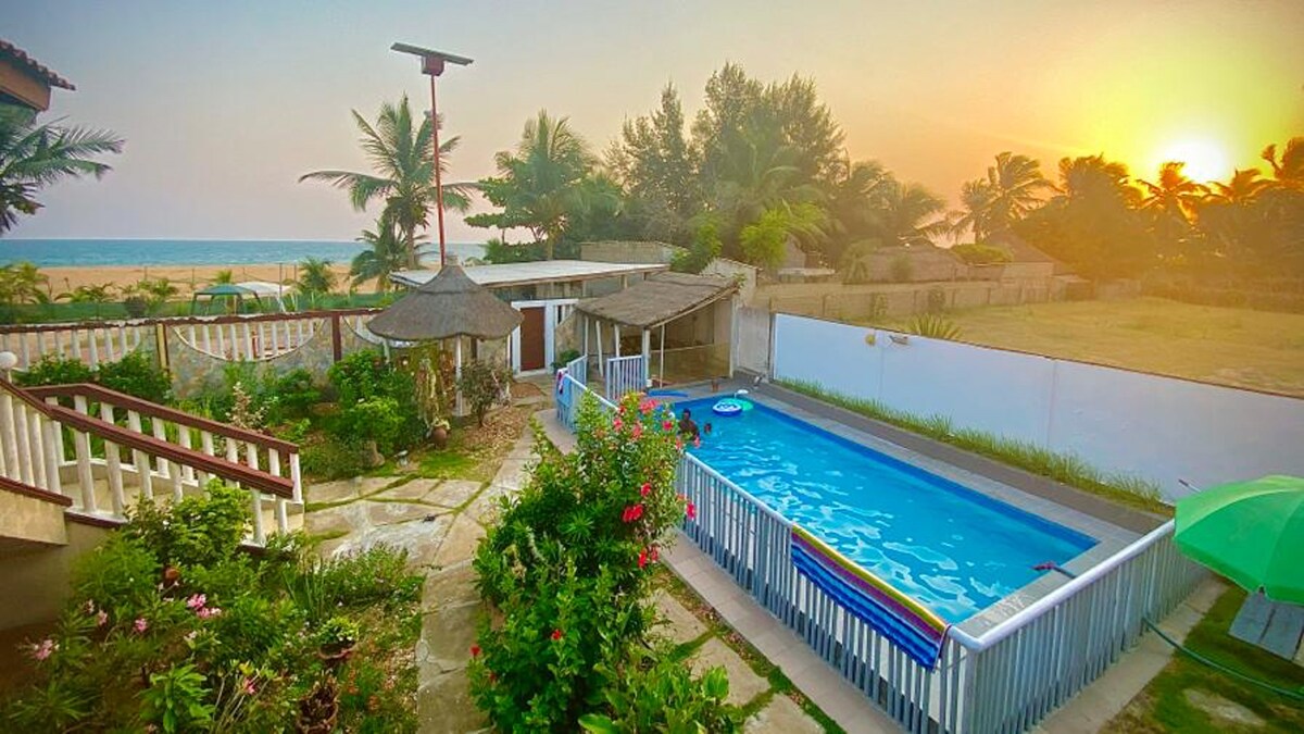 A shared pool is surrounded by vibrant gardens and palm trees, providing a refreshing retreat. The sunset casts a warm glow over the area, while the sea is visible in the background. Comfortable outdoor seating is situated near the pool, inviting relaxation.