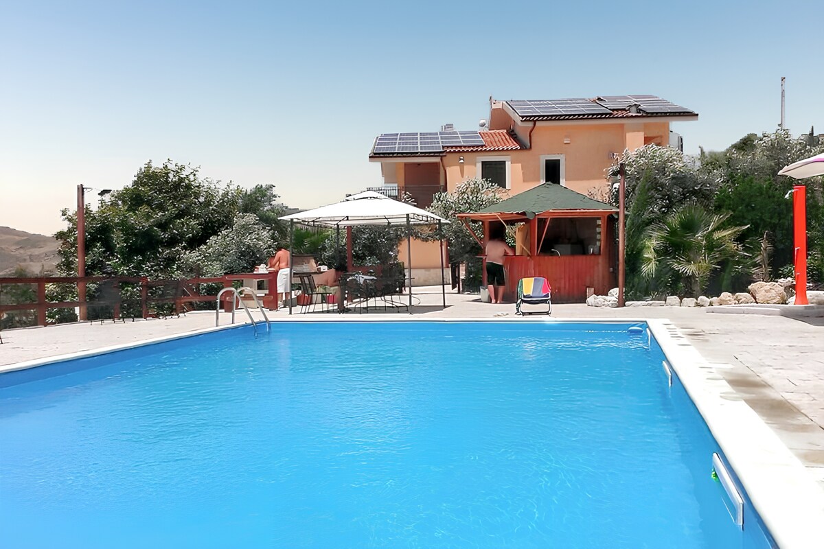A large private pool reflects the clear blue sky, accompanied by a spacious deck featuring sun loungers and shaded seating areas. The villa's exterior is visible in the background, surrounded by lush greenery and outdoor amenities, creating an inviting space for relaxation.