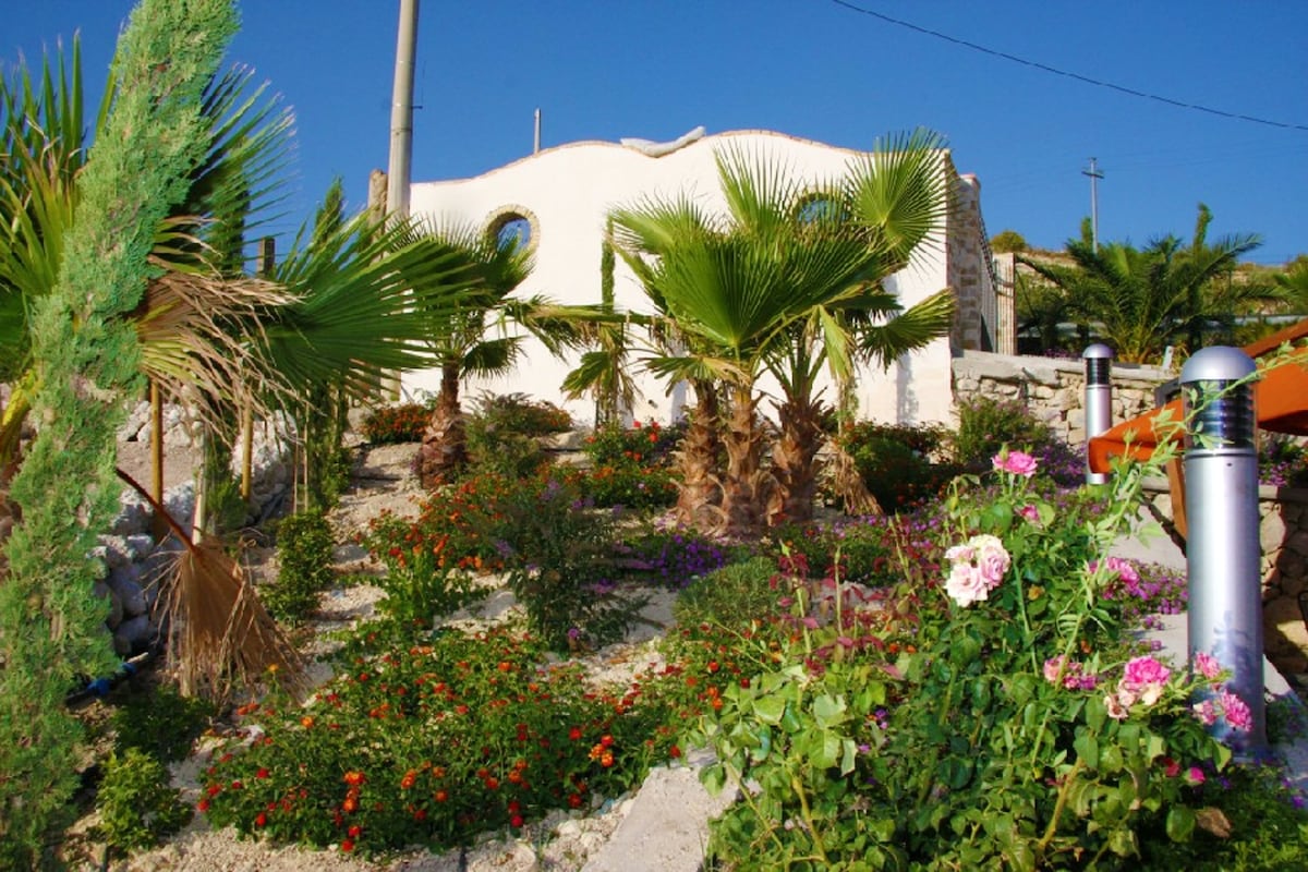 A vibrant garden surrounds the villa, featuring a variety of palm trees and colorful flowers. The well-maintained landscape adds a touch of nature, with blooming plants harmonizing with the architecture of the villa in the background.