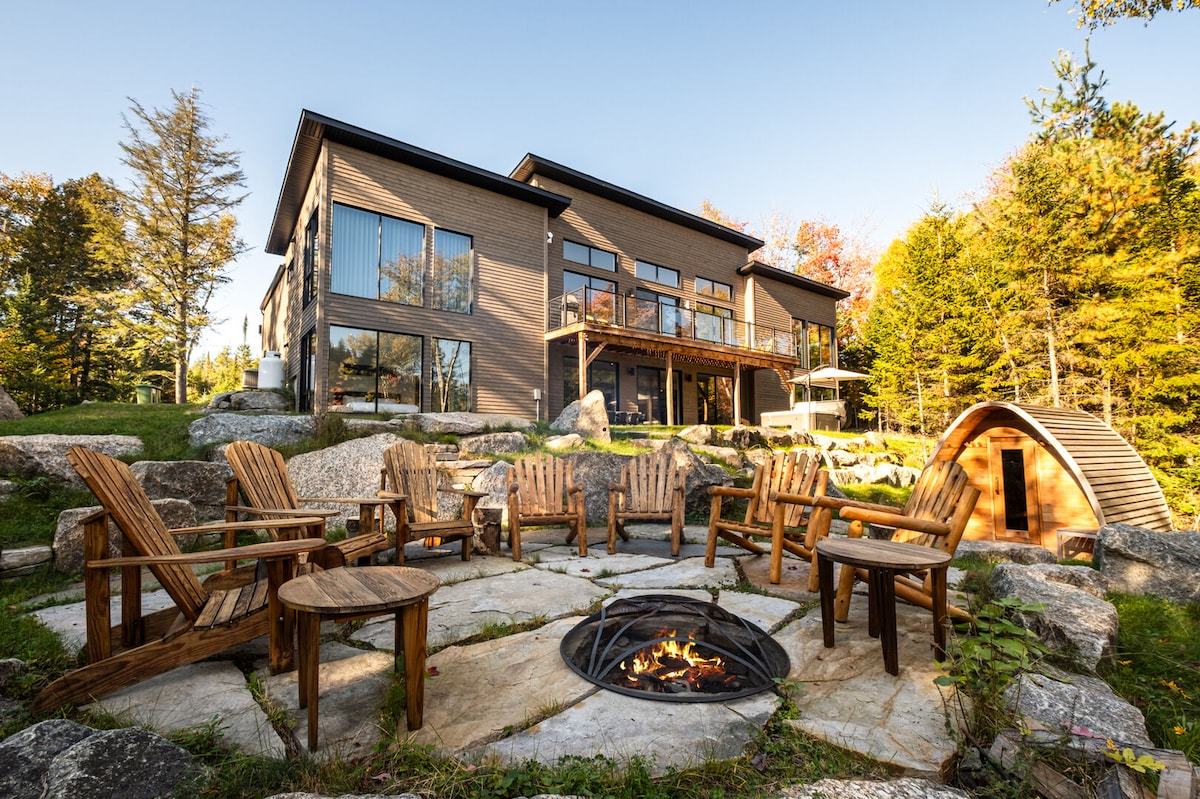 An outdoor seating area features rustic wooden chairs arranged in a circle around a fire pit. The modern house, with large windows, is set against a backdrop of trees, creating a peaceful environment. A wooden bridge leads to additional outdoor space.