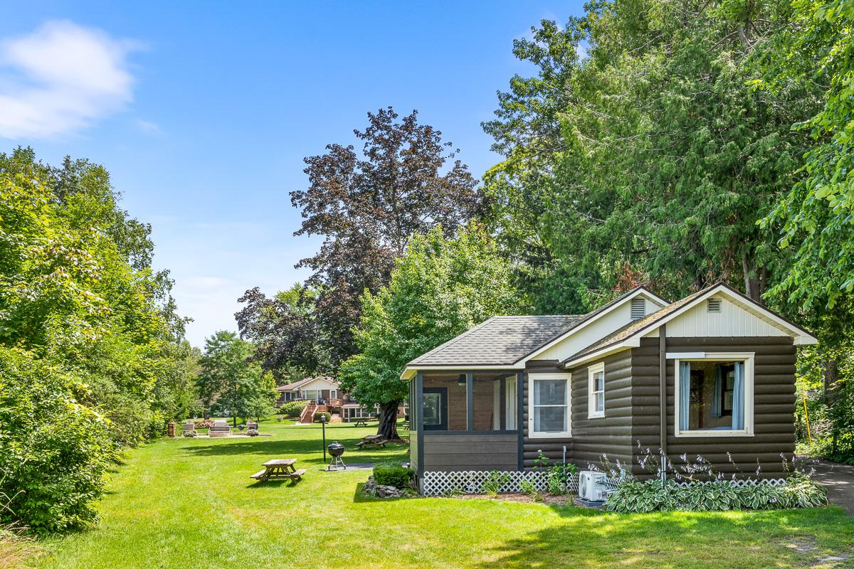 A log cabin sits amid a green landscape, framed by lush trees and open space. The cabin features a welcoming porch with large windows. A picnic area with wooden tables and a grill is visible nearby, providing a perfect setting for outdoor gatherings.