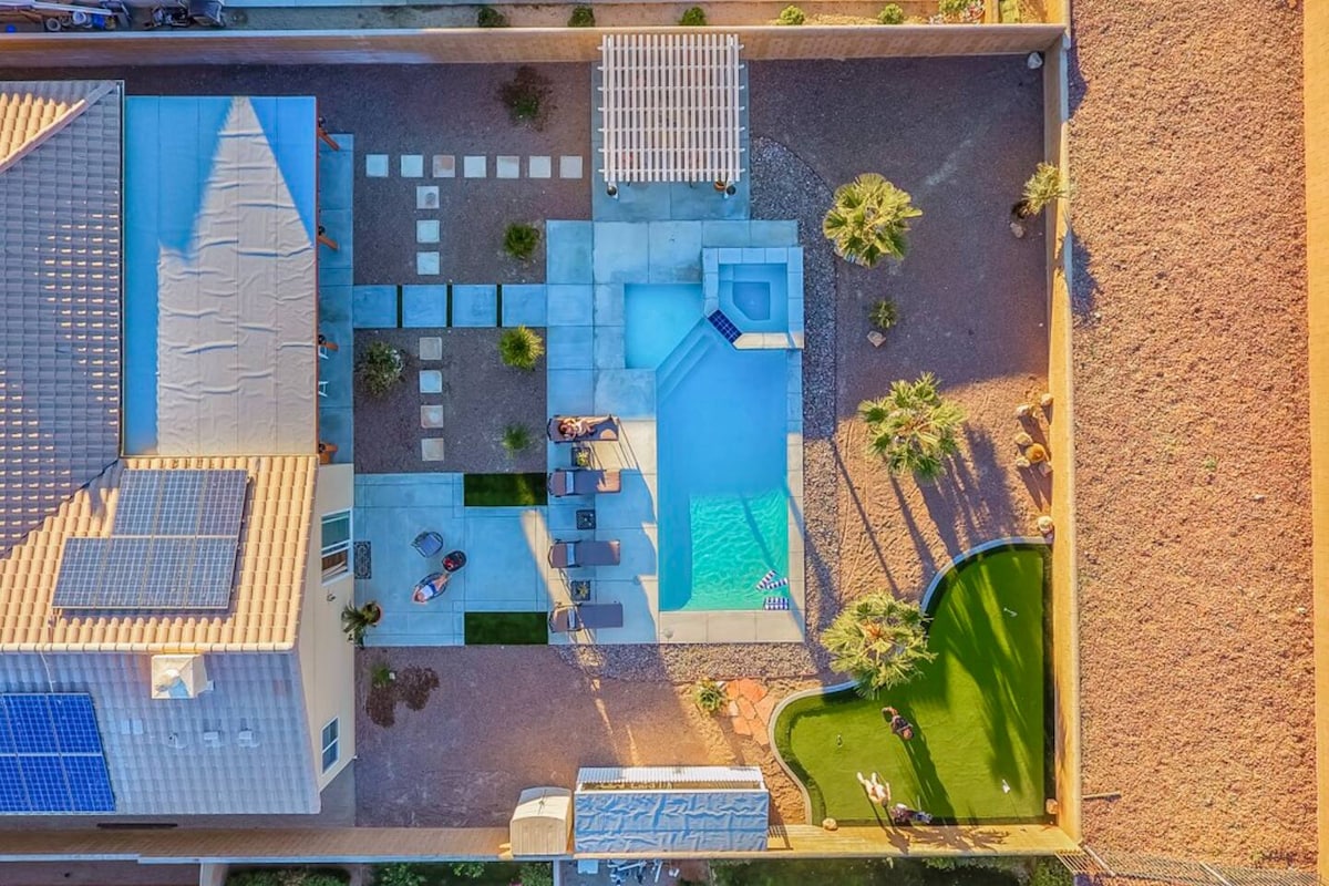 An aerial view of the villa's backyard features a private heated saltwater pool adjoining a hot tub. A spacious patio with lounge chairs and a BBQ grill can be seen, along with a putting green area. Desert landscaping and palm trees surround the space.