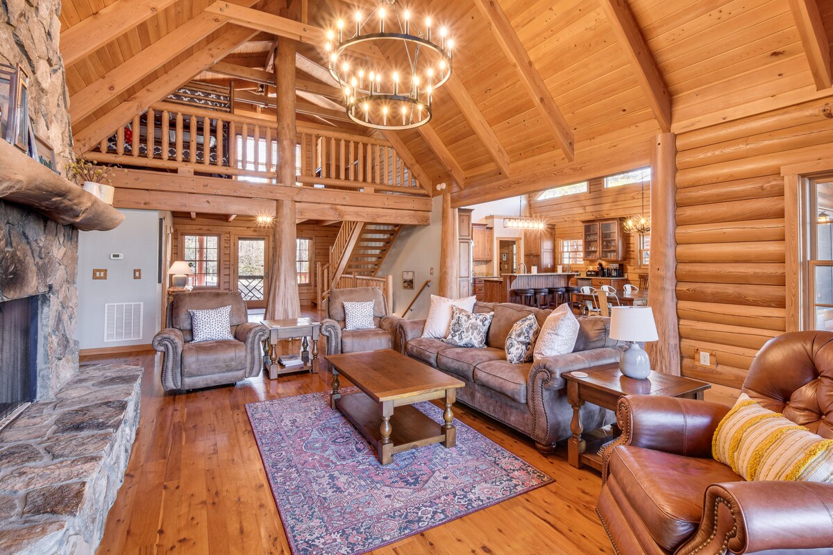 A spacious living area features high wooden beams and a stone fireplace. Comfortable seating arrangements include a leather armchair and a plush sofa adorned with decorative pillows. A large wooden coffee table sits at the center, complemented by a colorful area rug and a striking chandelier above.