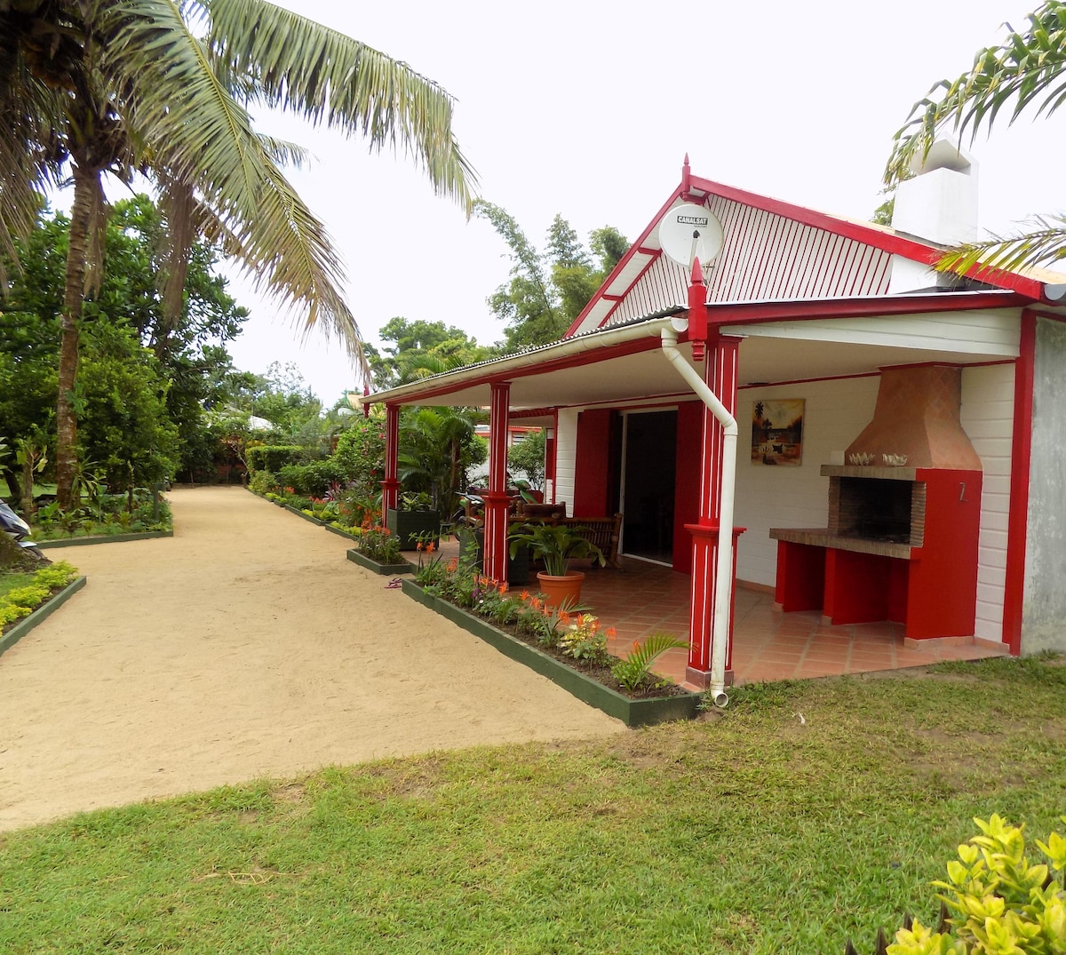 The studio is nestled in a vibrant garden, surrounded by tropical plants and palm trees. A covered terrace features outdoor seating and a stone barbecue, with a sandy pathway leading to the entrance. The bright red trim of the house adds a cheerful touch to the setting.