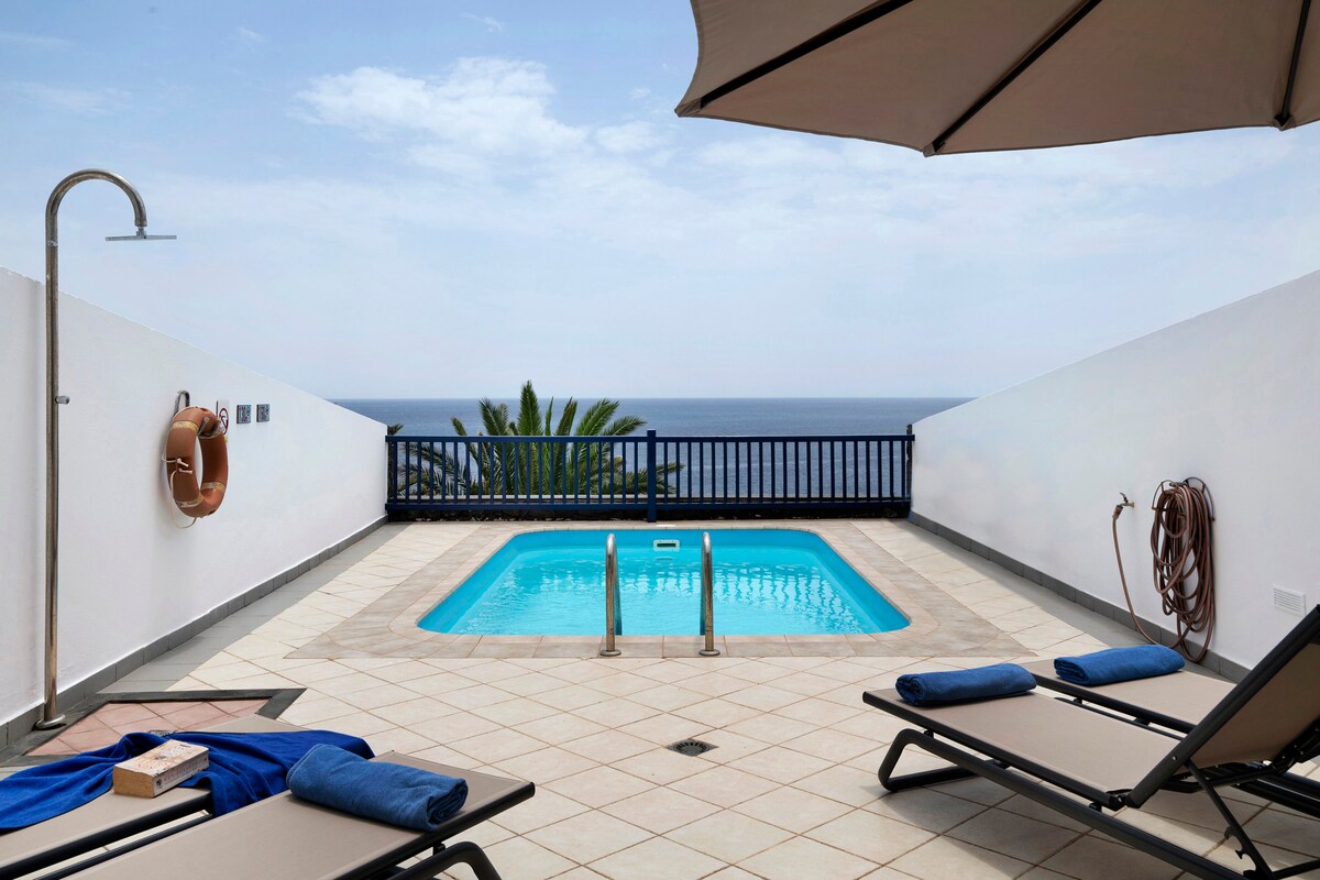 A private terrace features a swimming pool framed by tiled flooring. Sun loungers are positioned nearby, offering a place to relax. A sunshade provides partial coverage, while a view of the sea can be observed in the distance.