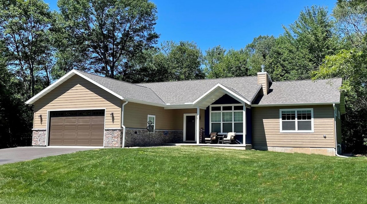 The exterior of the home is showcased, featuring a blend of stone and wood siding. A well-maintained lawn surrounds the entryway, which includes a seating area and a garage with a decorative door. Tall trees in the background enhance the serene setting.