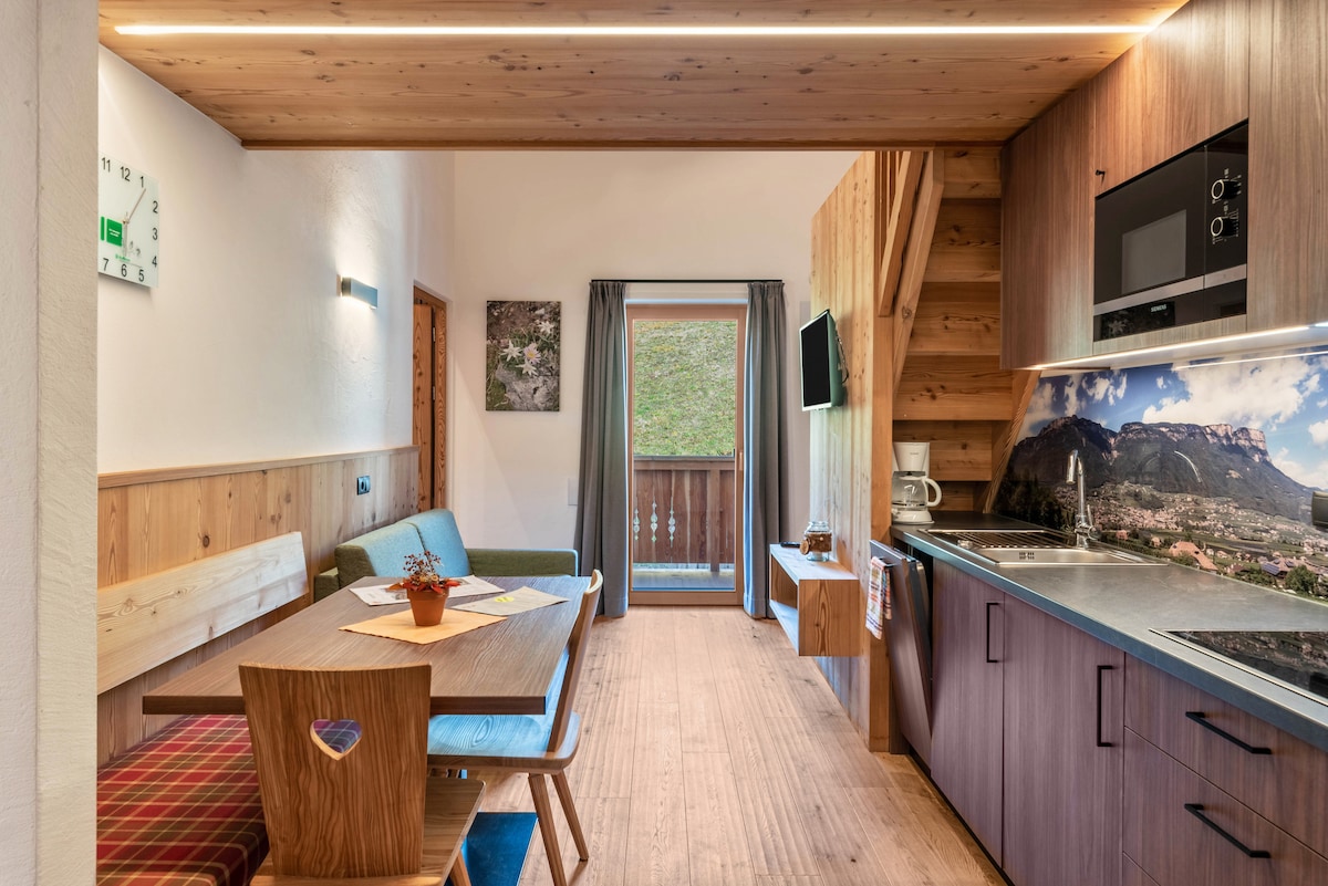 The living area features a well-equipped kitchen and a dining space with a wooden table and chairs. Natural light enters through the glass door leading to the balcony, which offers views of the surrounding landscape. A comfortable sofa is positioned against the wall.