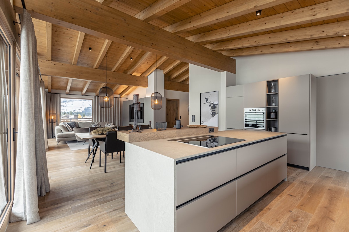 The open kitchen showcases modern cabinetry and a large central island with an induction cooktop. Wood beams highlight the ceiling, and large windows allow natural light to fill the space. Comfortable seating is visible in the adjacent living area.