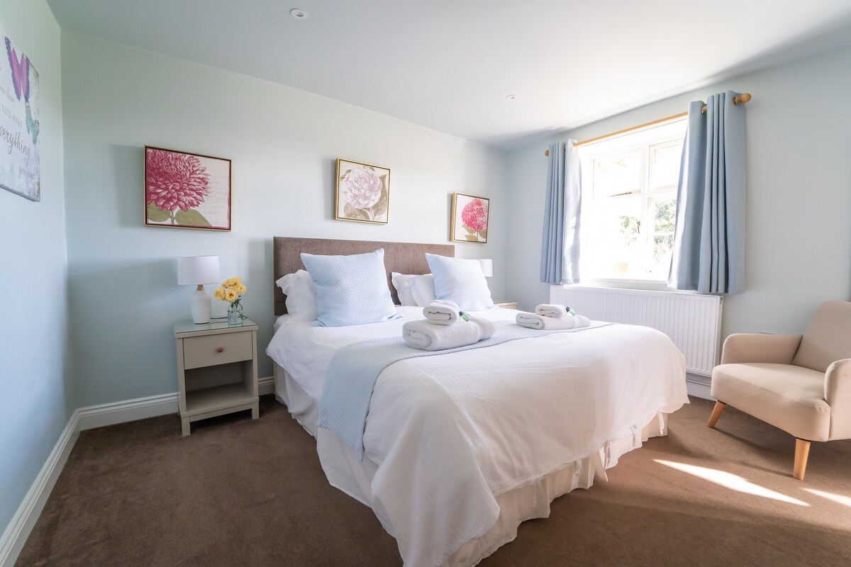 A serene bedroom features a spacious bed with crisp white linens and soft blue accents. Two decorative floral prints adorn the walls. The room is complemented by a cozy armchair and a bedside table with a vase of flowers. Natural light streams in through the window, partially covered by light curtains.