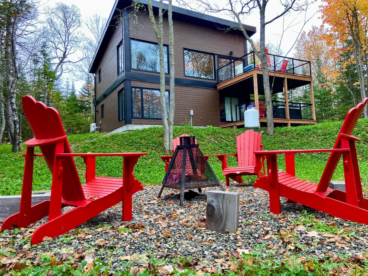 Four red Adirondack chairs are arranged around a small fire pit on gravel, creating an inviting outdoor seating area. The chalet, with large glass windows, is visible in the background, surrounded by trees with autumn foliage.