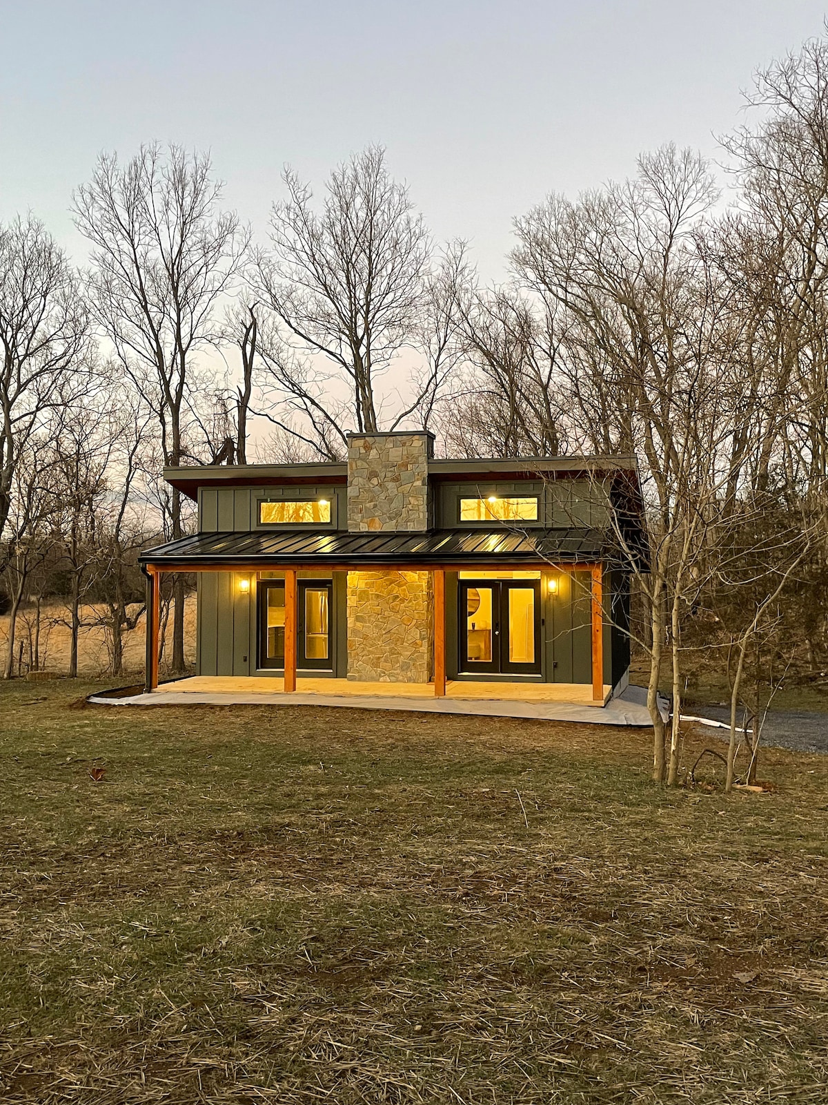 The exterior of a two-story cottage is presented, featuring a blend of dark green siding and stone accents. Large windows are illuminated, highlighting a welcoming entrance. Surrounding trees are bare, indicating the season, while a spacious yard of grass and earth is visible in the foreground.