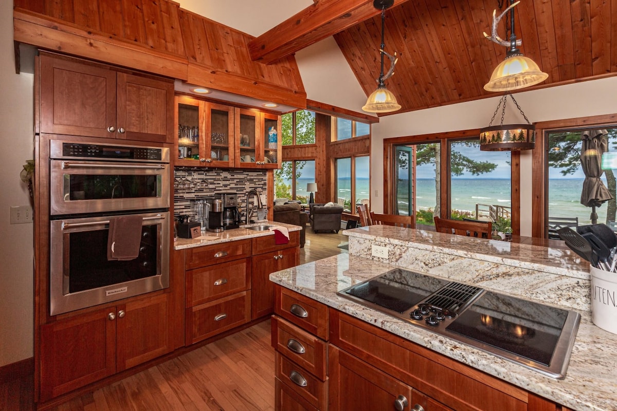 A spacious kitchen features warm wood cabinetry and a large granite island. Modern stainless steel appliances are integrated, with a scenic view of Lake Superior visible through expansive windows. The open-concept design connects the kitchen to a cozy living area, enhancing the cabin's inviting atmosphere.