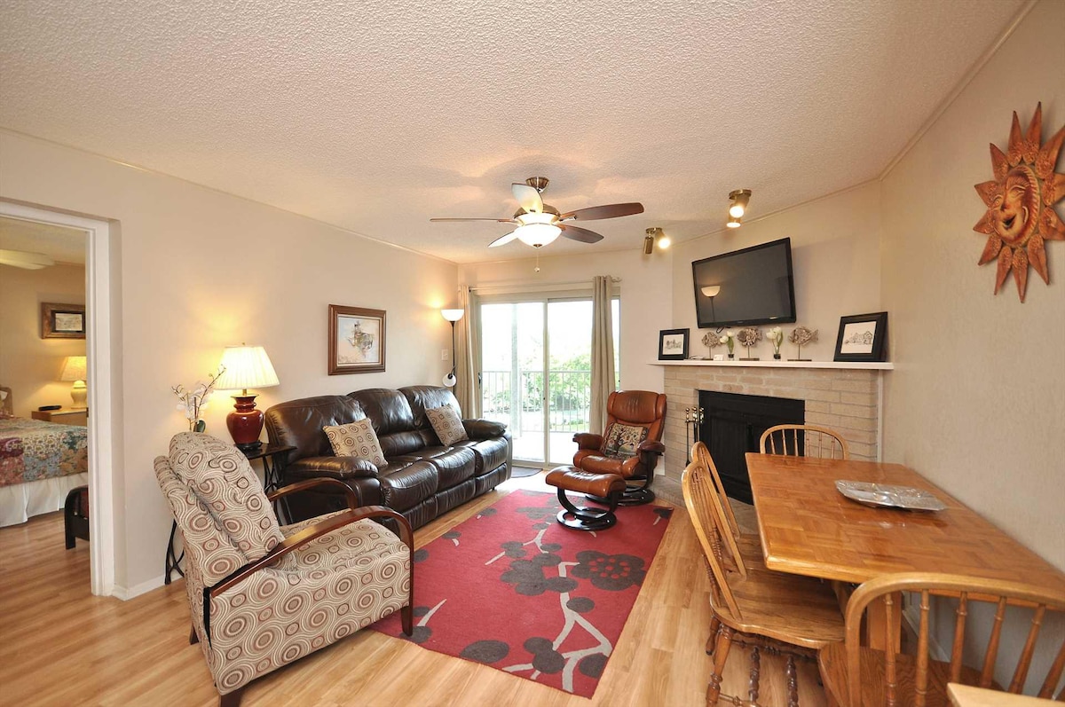 A comfortable living area is presented, featuring a brown leather sofa and armchair arranged on a patterned area rug. A wooden dining table with seating for four is visible. A ceiling fan hangs above, and natural light enters through sliding glass doors that lead to the balcony.