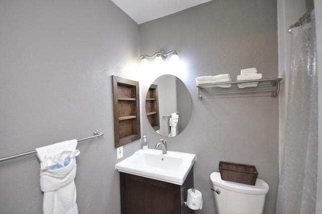A modern bathroom features a circular mirror above a clean sink with a dark vanity. Shelves hold neatly arranged towels, and a wicker basket adds a touch of warmth. A toilet is positioned near the wall, enhancing the space's functionality.