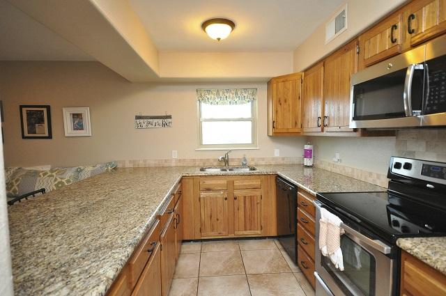 A well-appointed kitchen features wooden cabinetry and a spacious granite countertop. Stainless steel appliances, including an oven, microwave, and dishwasher, are visible. Natural light enters through a nearby window adorned with a patterned valance, enhancing the functional layout of the kitchen space.