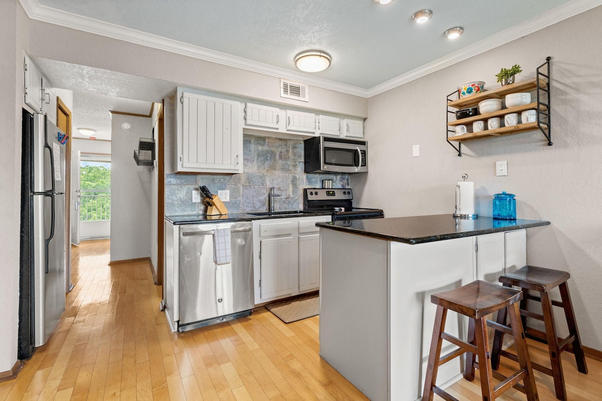 A modern kitchen is equipped with stainless steel appliances and a black countertop. Wooden cabinets display dishware, and a small dining area features two wooden stools. Light wood flooring enhances the spacious feel, and a doorway leads to a bright, airy living area.