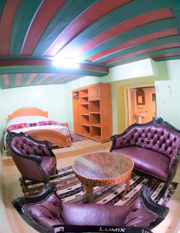 A cozy room with a spacious bed is complemented by two dark leather armchairs surrounding a circular wooden table. Textured, green beams adorn the ceiling, while a wooden shelving unit provides storage. A warm rug adds comfort to the wooden flooring.