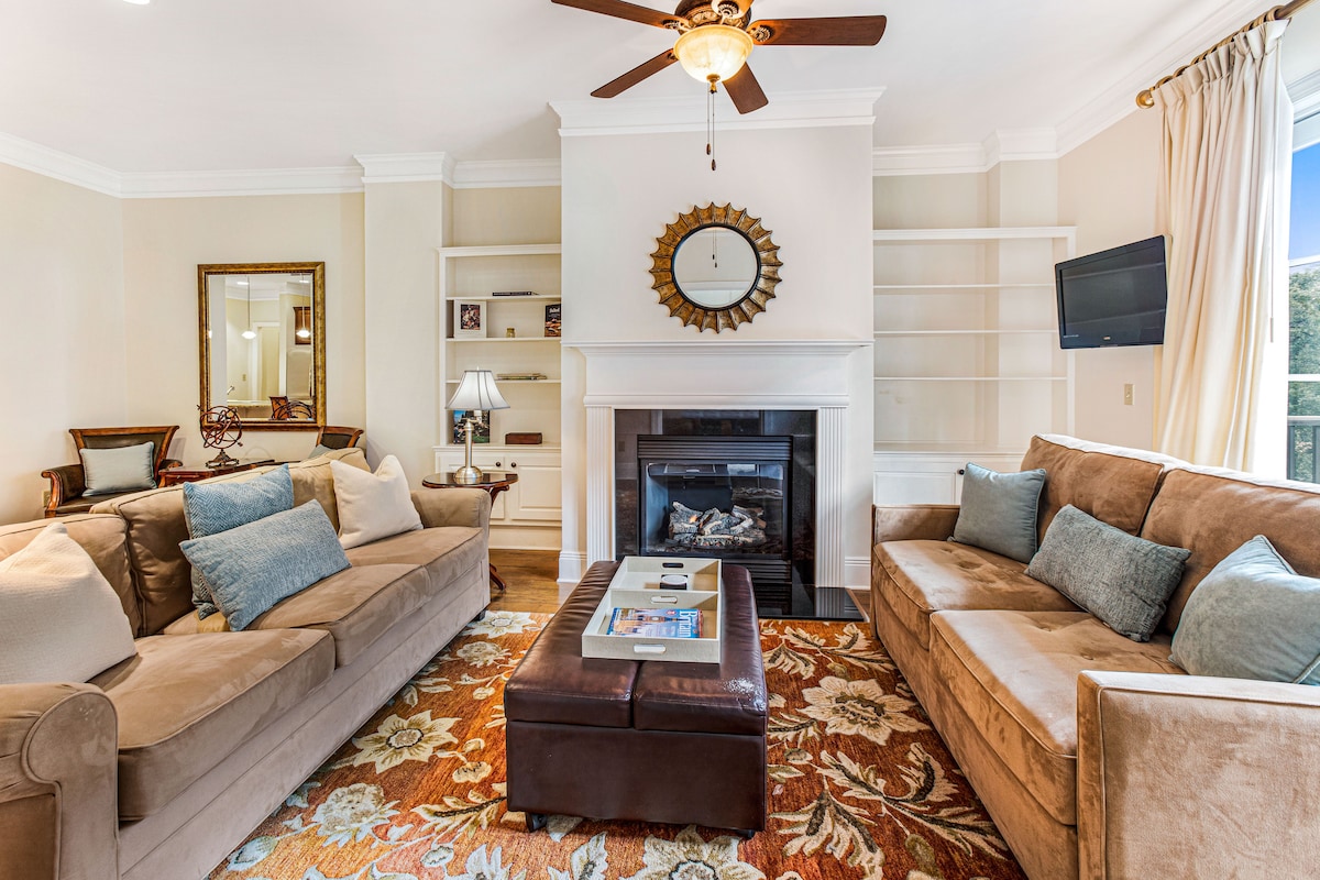 The living space features two plush sofas arranged around a coffee table, with a decorative area rug beneath. A fireplace is set in the wall, complemented by built-in shelves. Natural light flows in through large windows, and a wall-mounted television is also present.