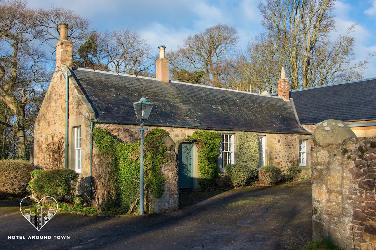 A charming cottage is framed by lush greenery and inviting pathways. Its stone exterior features a traditional roof and subtle architectural details. Windows provide glimpses of the interior, while a rustic lantern adds character to the entrance. The surrounding landscape is dotted with trees, enhancing the serene setting.