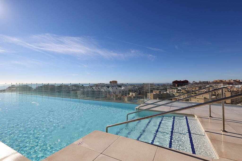An inviting infinity pool extends to the horizon, providing stunning views of the sea and city. The pool's clear water reflects the blue sky, while smooth deck tiles surround it. The area includes accessibility features, ensuring ease of access for all guests.