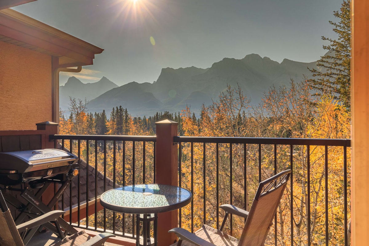 A private balcony is featured, furnished with a round table and two chairs. A grill is positioned nearby. The backdrop showcases a scenic view of mountains bathed in sunlight, with vibrant autumn foliage adorning the landscape.