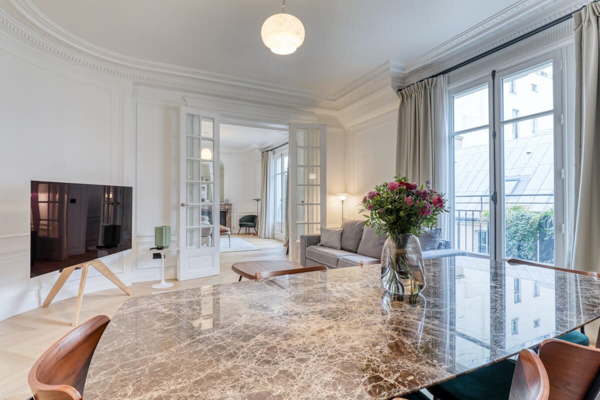 A spacious living area features a marble dining table surrounded by wooden chairs. A gray sofa is positioned near a large window, which is dressed with soft curtains. A flat-screen TV stands on a minimalist wooden stand, with a vase of fresh flowers adding a touch of color.