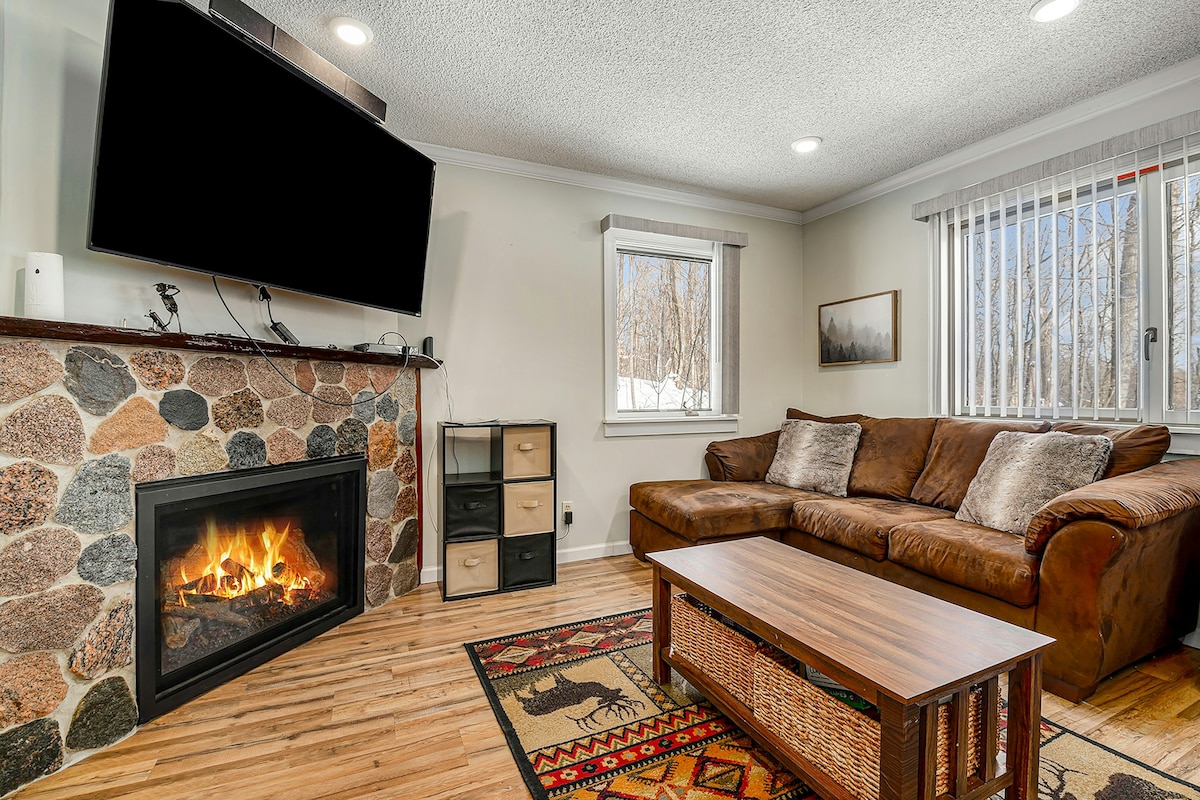 A spacious living area features a stone fireplace and a comfortable brown leather sectional sofa. A wooden coffee table rests on a patterned rug with natural light filtering in through the window, enhancing the cozy atmosphere.