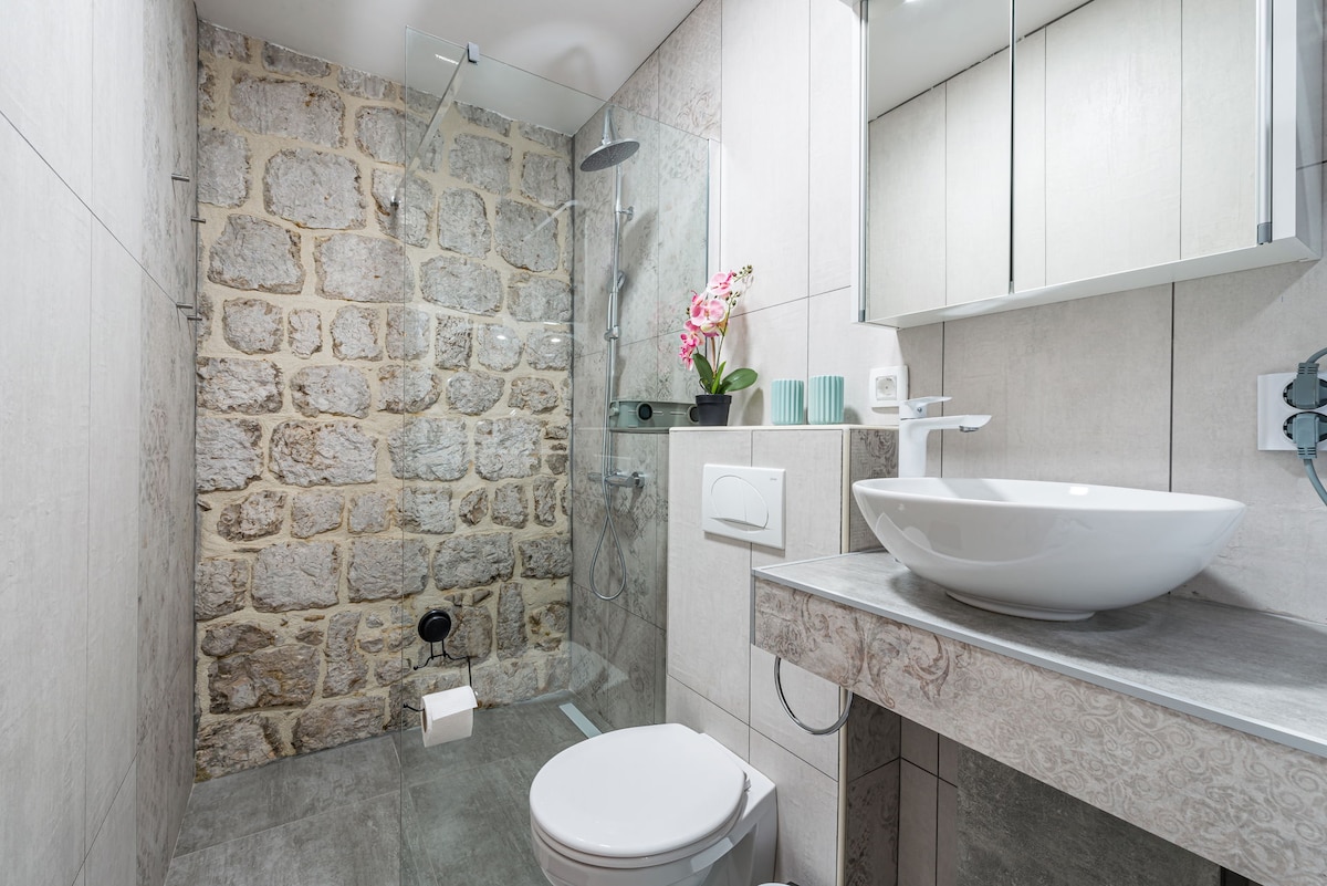 A modern bathroom showcases a stylish design with natural stone walls. A sleek, round sink is positioned on a light gray countertop. A glass shower is equipped with a rain showerhead, while a toilet is discreetly placed. Decorative plants and lavender accents add a touch of color.