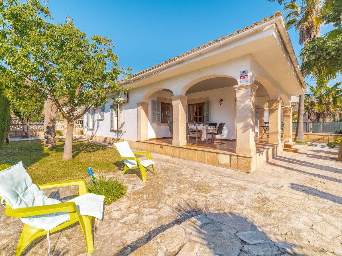 A sunlit patio features stone flooring and arched columns, providing a shaded area. Two bright green lounge chairs are positioned for relaxation, while a wooden dining table is set for outdoor meals. Lush trees add greenery to the surrounding space.