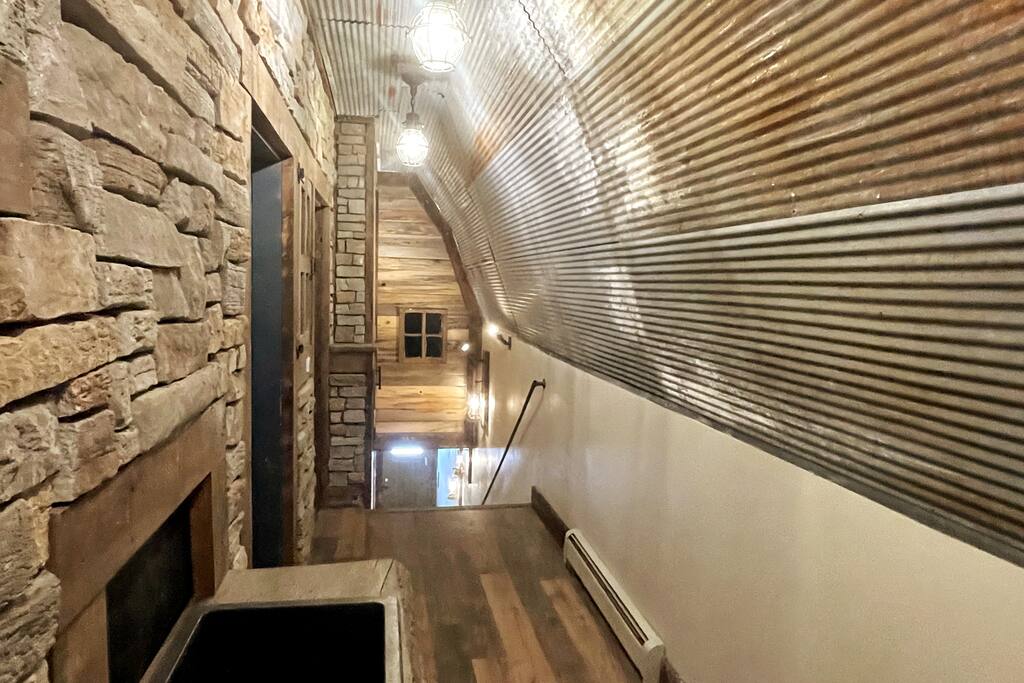 A stylish staircase is framed by rustic stone walls and corrugated metal, leading to an inviting loft space. Soft lighting from pendant fixtures enhances the warm wood accents, while the smooth wood flooring adds a natural touch to the environment.