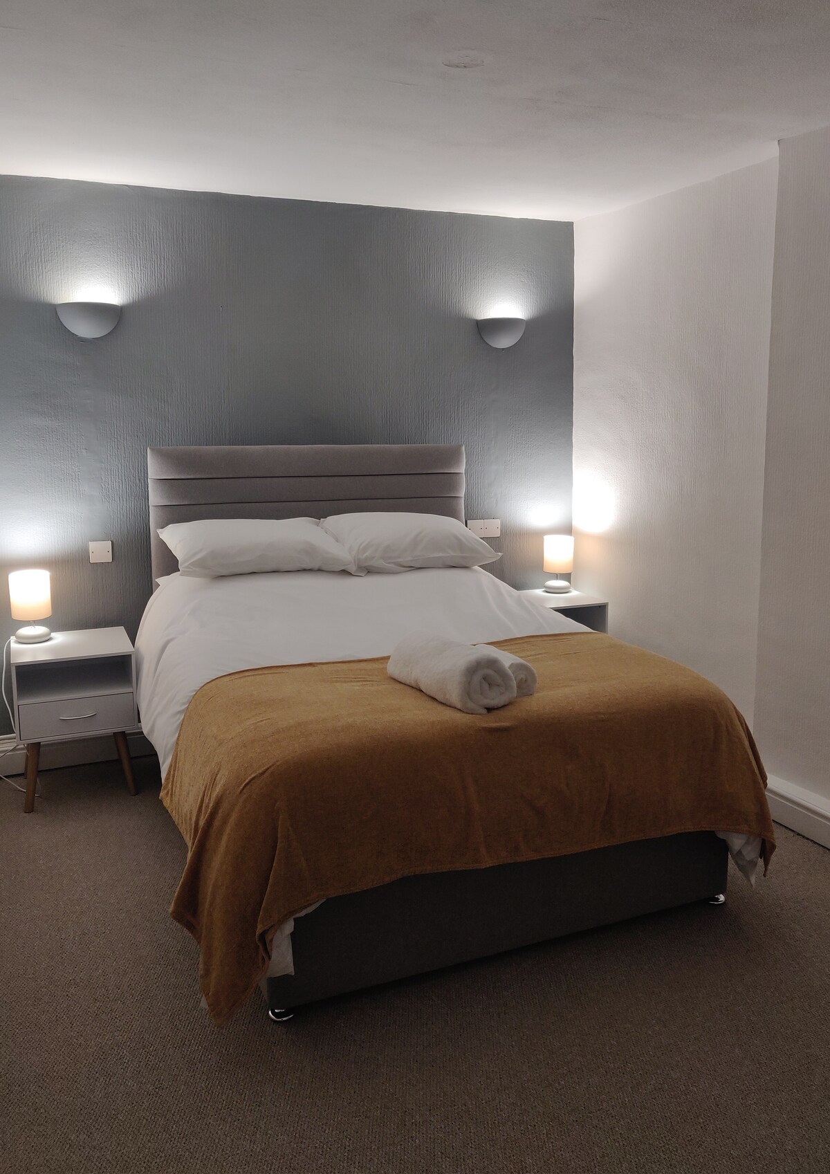 A newly refurbished double room features a comfortable bed with a soft blanket and a towel elegantly placed on top. Soft wall lighting illuminates the space, accentuating the neutral color scheme and inviting ambiance of the room.