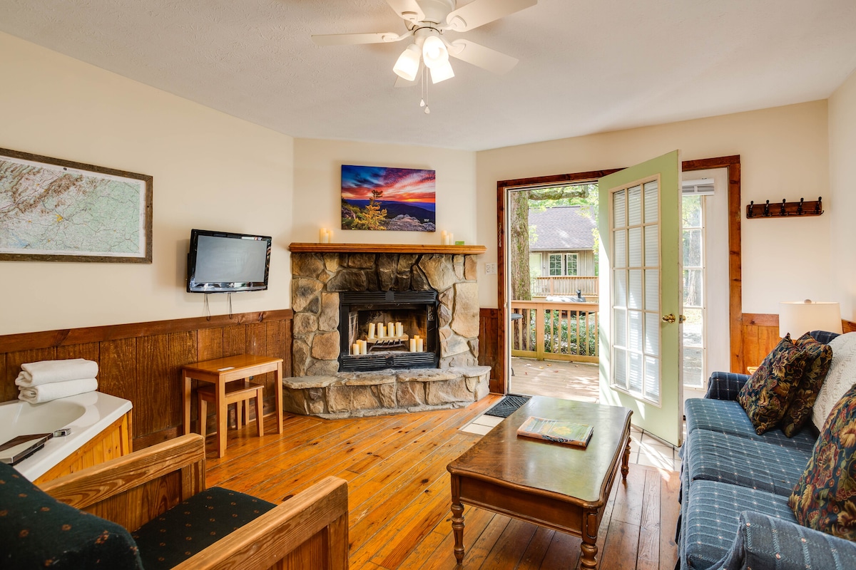 A cozy living area features a stone fireplace with a decorative mantle. A large window opens to the outdoor deck, allowing natural light to fill the space. Comfortable seating arrangements include a blue sofa and wooden chairs. A TV is mounted on the wall opposite the fireplace.