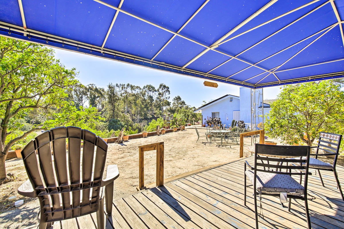 An outdoor patio area is shaded by a blue canopy, featuring a wooden deck with black chairs and a table. Surrounding greenery and potted plants create a natural setting, with a path leading towards a white building in the distance.