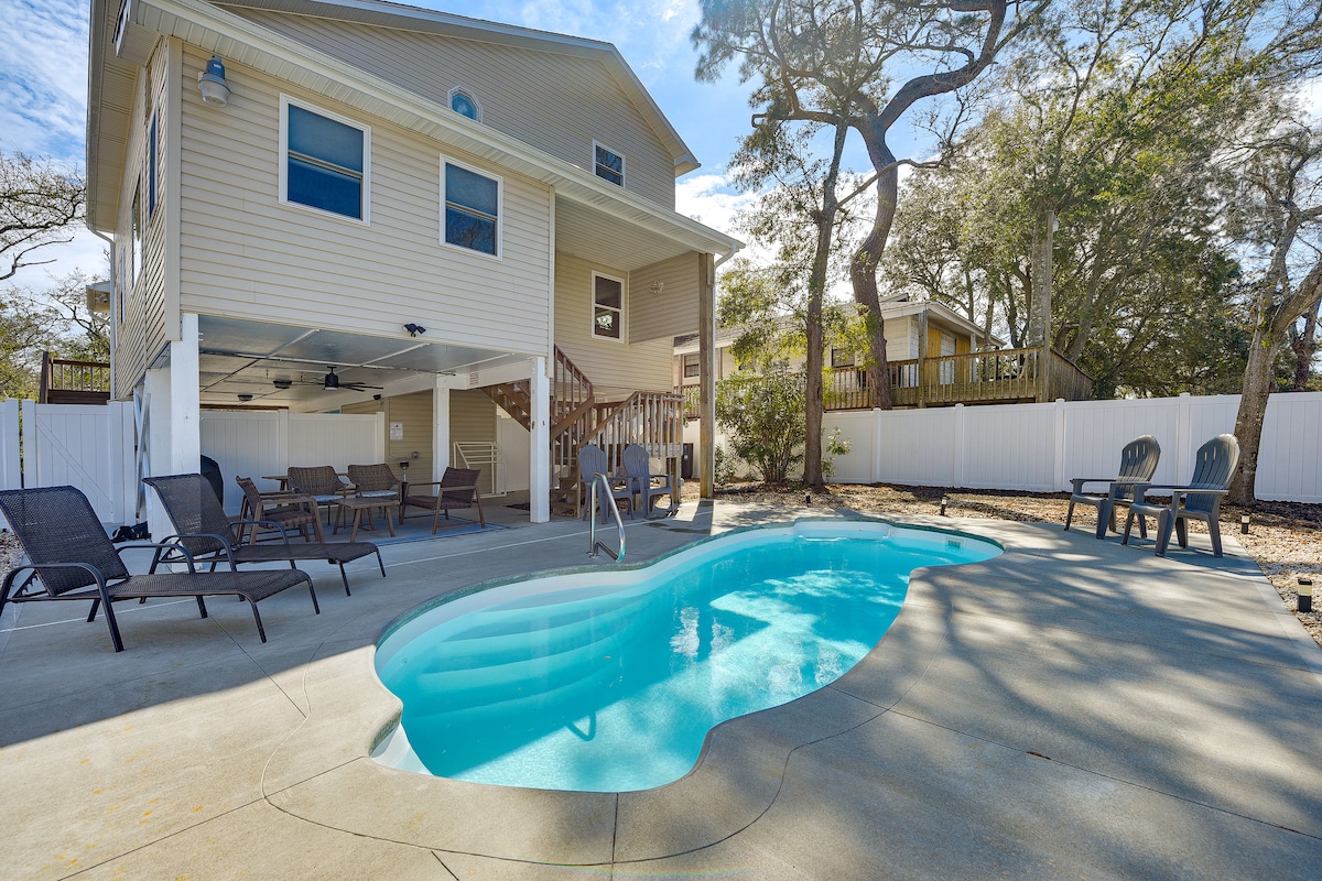 An inviting backyard features a private, heated pool with a gentle curve and steps leading into the water. Sun loungers are positioned around the pool area, and a covered patio provides shade with comfortable seating. Lush trees and fencing create a sense of privacy.